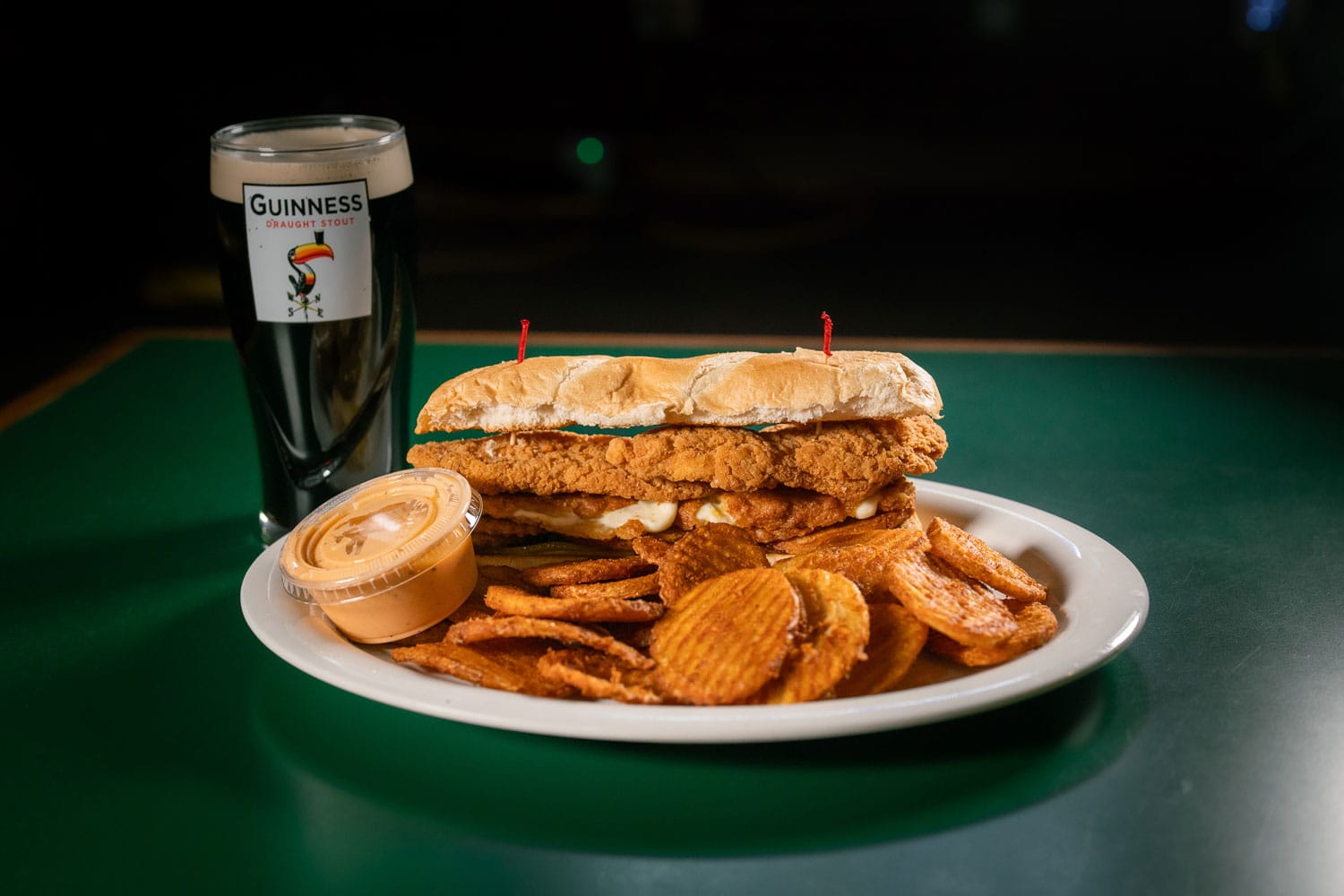 White plate with a fried chicken sandwich, potato chips, and a small cup of dipping sauce; a Guinness stout glass sits behind on a dark table.